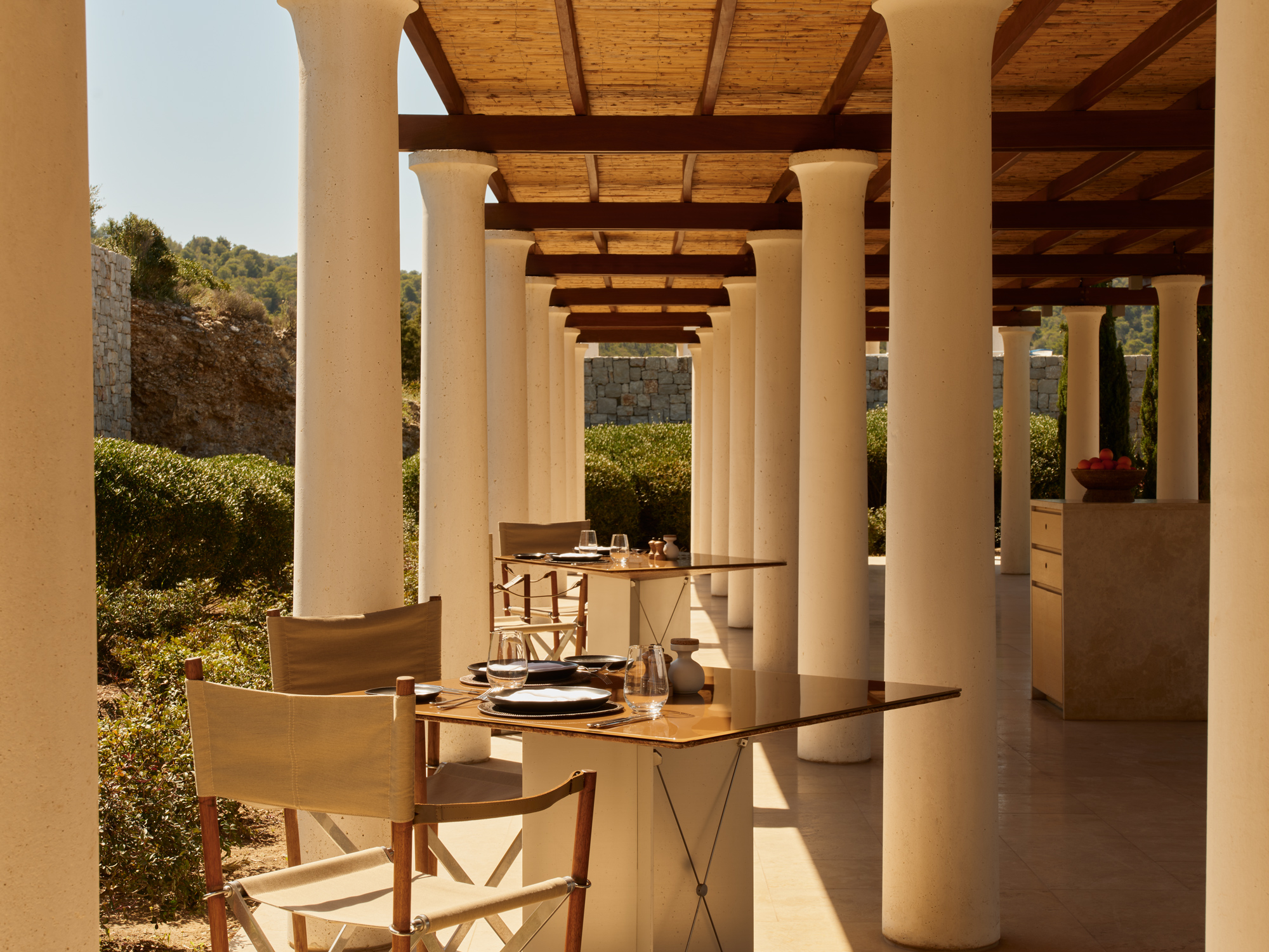 Elegant outdoor dining area at Amanzoe, featuring wooden tables and chairs under a colonnade with lush greenery in the bac...