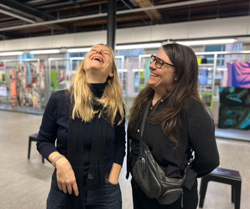 Two women laughing together in an art gallery, surrounded by colorful woven artworks.