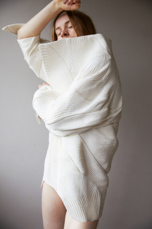 Model wearing a textured, oversized white garment with a sculptural design, posed against a neutral background.