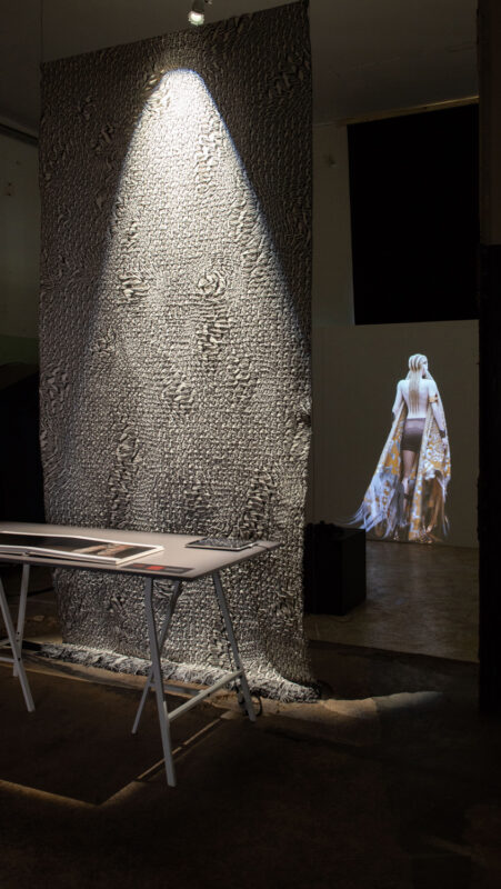 Textured fabric wall illuminated by a spotlight, with a figure in a flowing garment projected in the background.