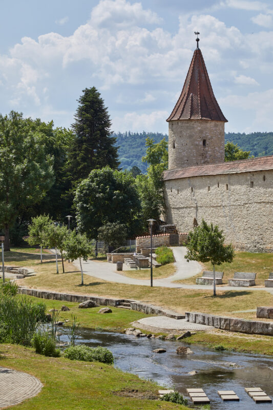 Historic stone tower with a red-tiled roof beside a grassy area and a stream, under a blue sky with clouds.