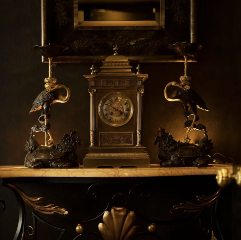 Elegant antique clock flanked by ornate candle holders with crane figures, set against a dark, luxurious background.