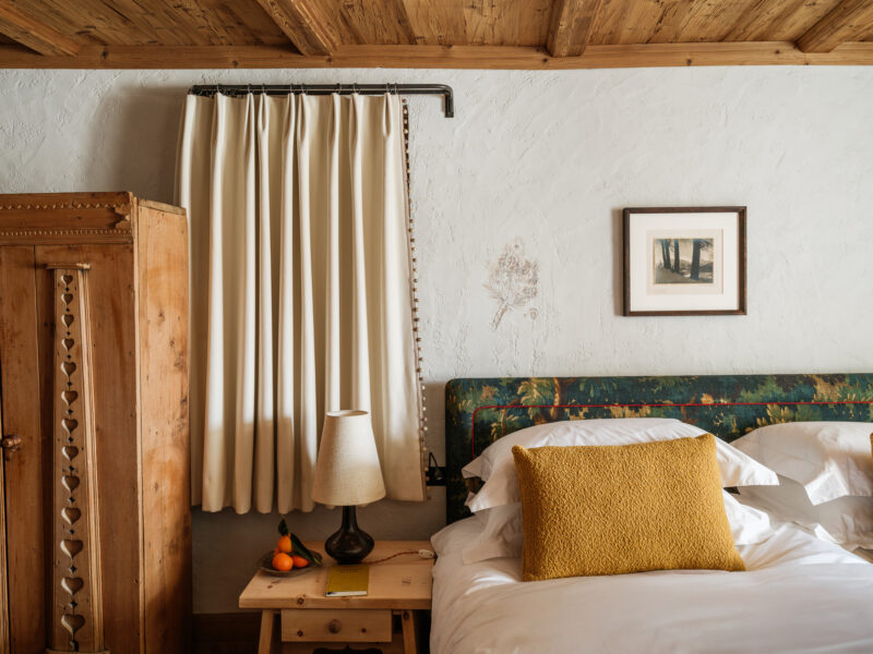 Cozy bedroom featuring a wooden bed with a yellow pillow, a nightstand with a lamp, and curtains beside a vintage wardrobe.