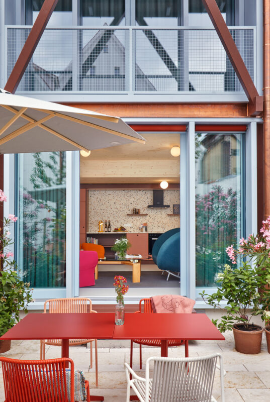 Modern outdoor seating area with a red table and chairs, overlooking a stylish interior of the Engelwirt Hotel.