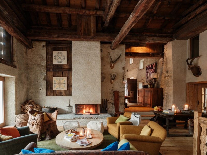 Cozy lounge area in Chesa Marchetta with a fireplace, colorful seating, and rustic decor featuring antlers and artwork.