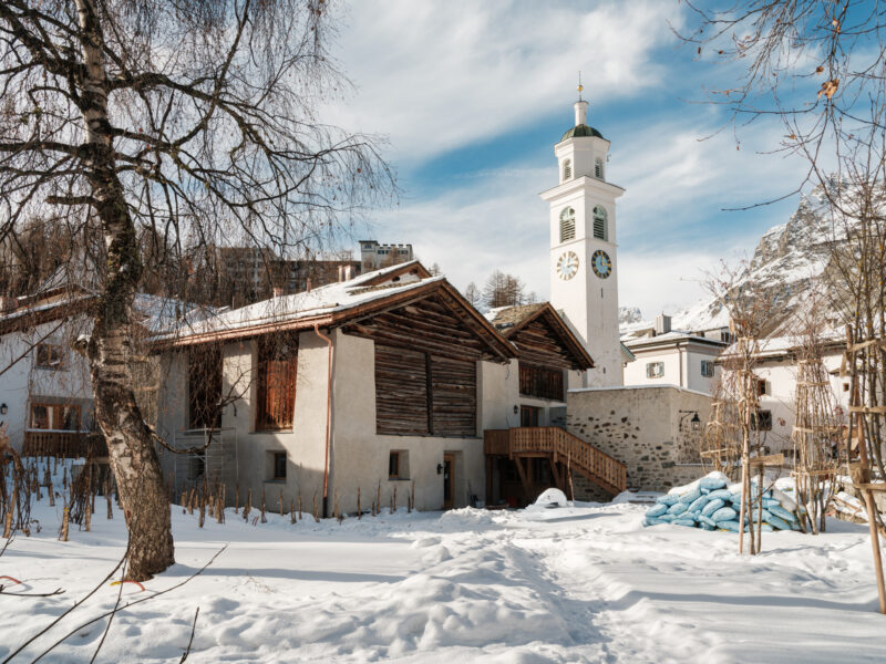 Snow-covered exterior of Chesa Marchetta restaurant, featuring a clock tower and surrounding buildings against a blue sky.