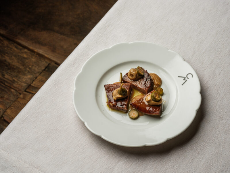 Veal tongue tonnata served on a white plate, garnished with capers and herbs, on a textured table surface.