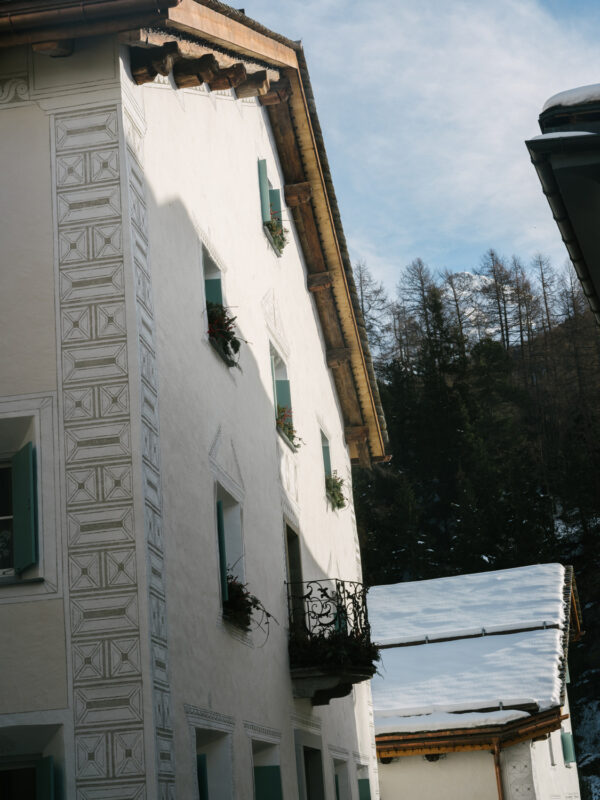 White building with decorative patterns and green shutters, featuring a balcony, surrounded by snow-covered roofs and trees.