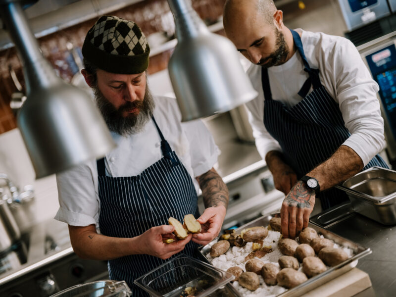 Two chefs in a kitchen preparing gnocchi, one peeling potatoes while the other arranges them on a tray.