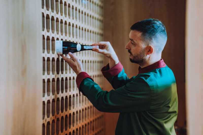 Man in a green and burgundy outfit placing a wine bottle into a wooden wine rack with circular openings.