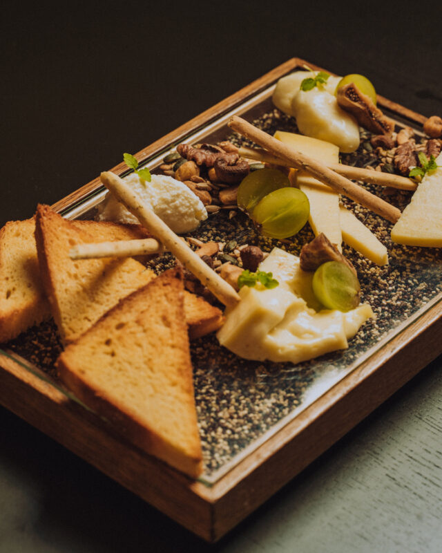 Cheese platter with various cheeses, grapes, nuts, and toasted bread on a wooden serving board.