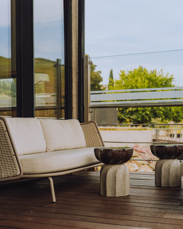 Modern balcony setting featuring a white sofa and two textured side tables, with greenery visible in the background.