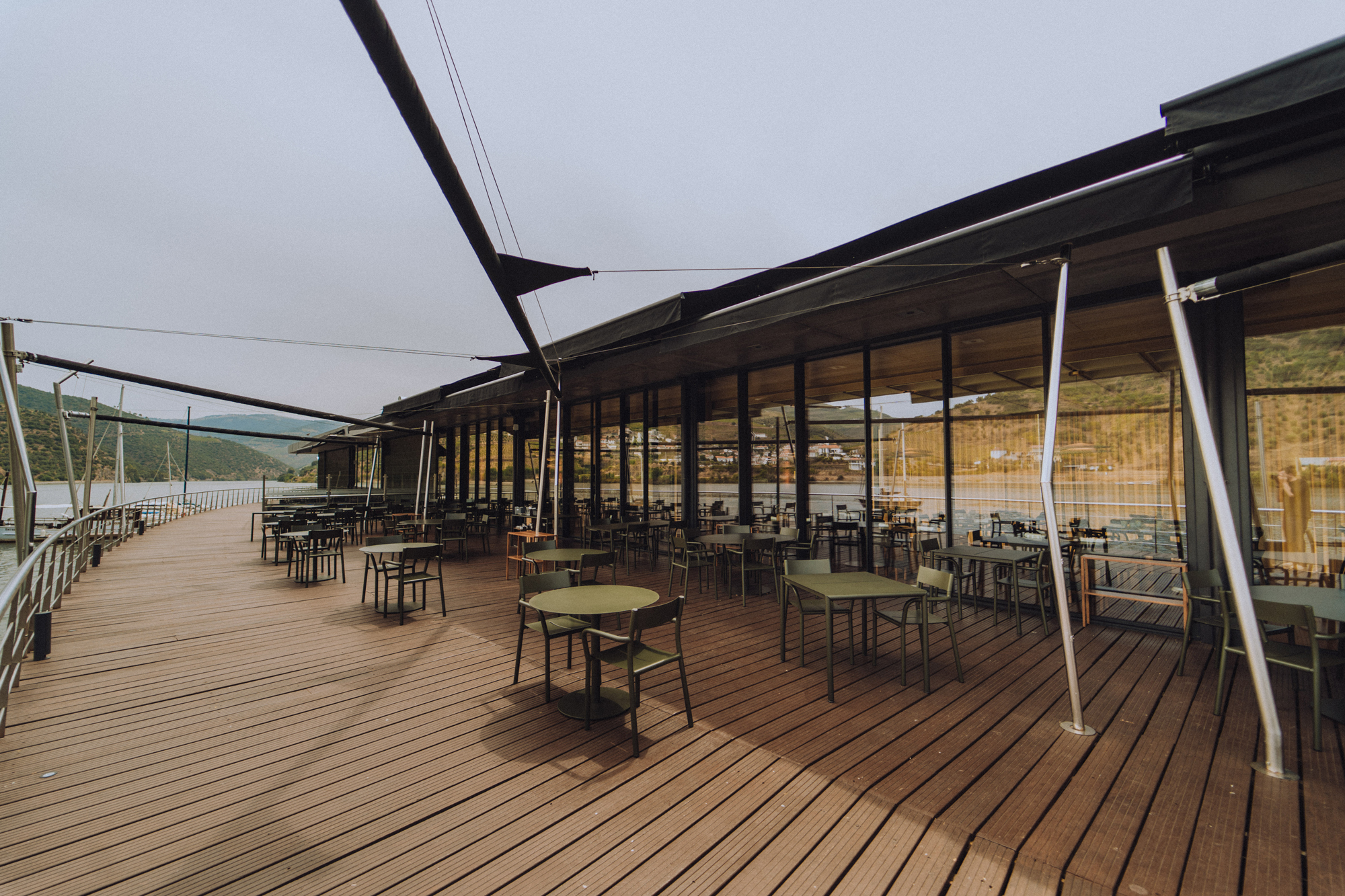 Modern restaurant terrace with empty tables overlooking a scenic view, surrounded by glass walls and wooden decking.