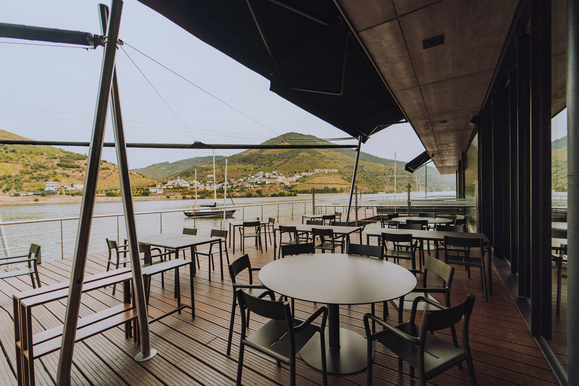 Modern outdoor dining area with tables and chairs overlooking a serene waterfront and hills in the background.
