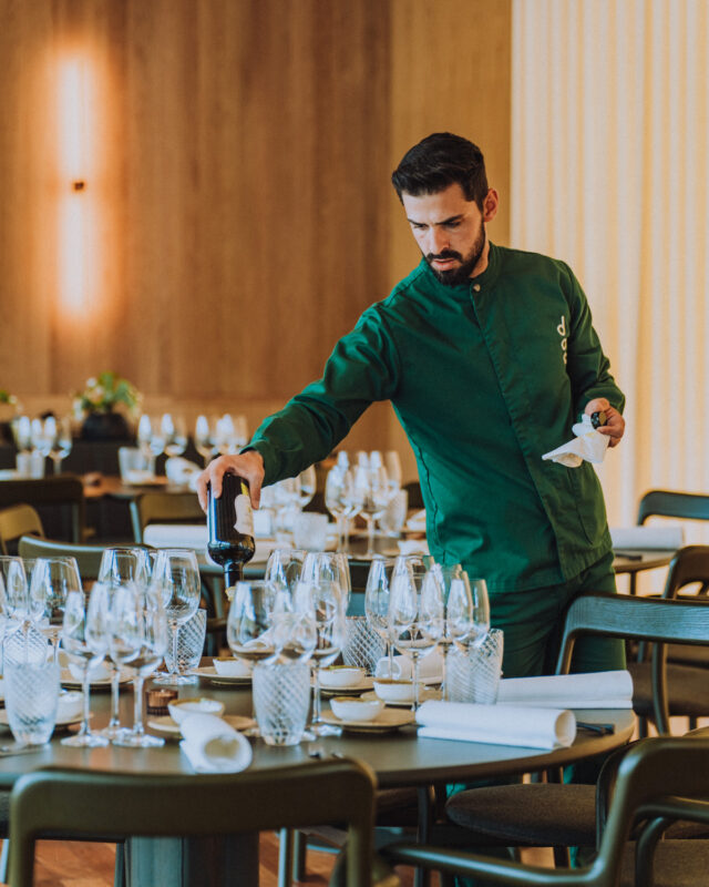 A waiter in a green jacket pours wine into a glass at a beautifully set dining table.