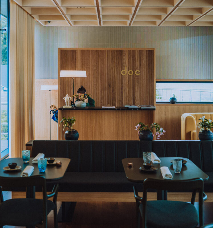 Modern café interior featuring a wooden counter with the word "doc," plants on tables, and a barista at work.