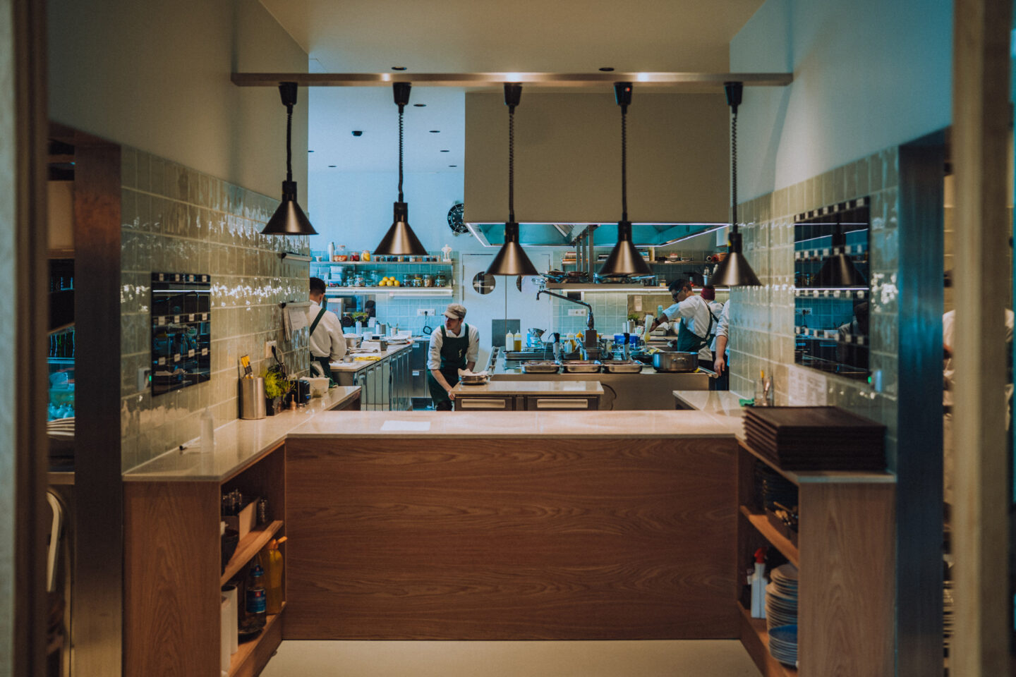 Modern kitchen interior with chefs working, featuring a wooden counter and pendant lighting.