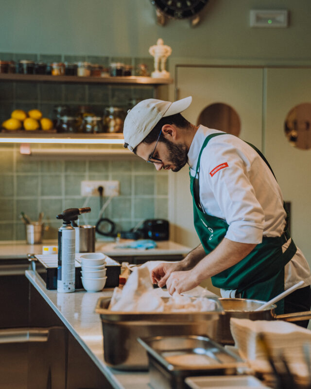 Chef in a green apron prepares food in a modern kitchen, focused on plating with kitchen tools and ingredients visible.