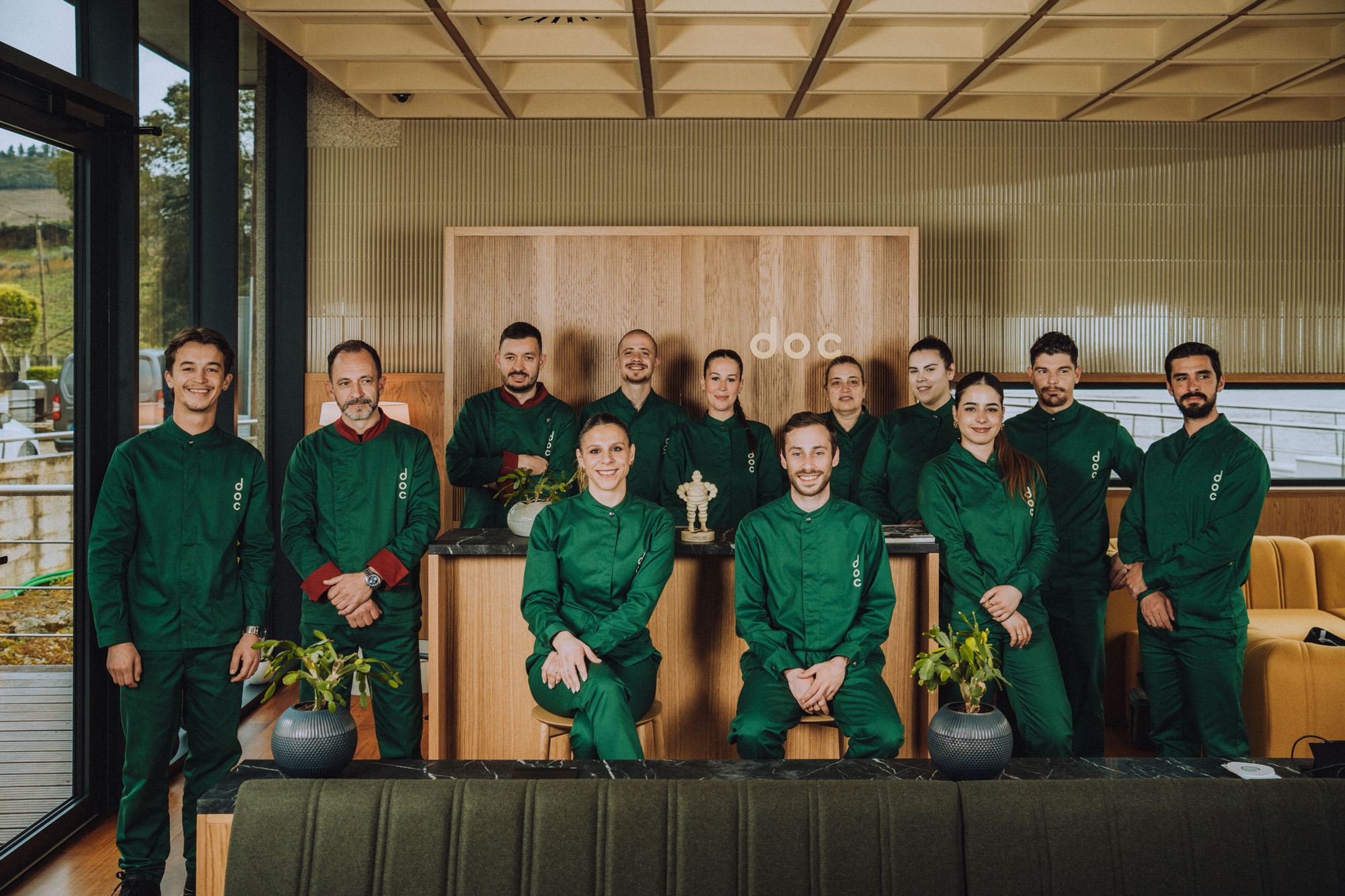 Group of twelve people in green uniforms posing together in a modern indoor setting with plants and a trophy.