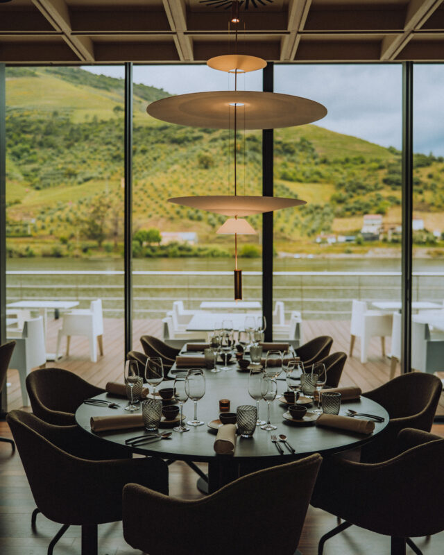 Elegant dining setup with a round table, glassware, and scenic river view through large windows.