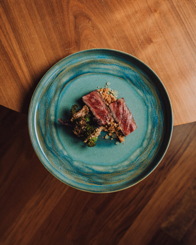 Plate with a portion of sliced meat atop a bed of grains and greens, set on a wooden table.