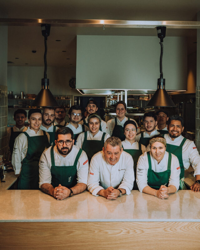 A group of chefs in green aprons poses together in a modern kitchen, smiling at the camera.