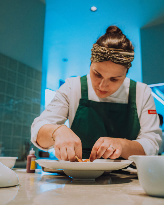 Chef carefully plating a dish in a modern kitchen, wearing a green apron and a patterned headband.