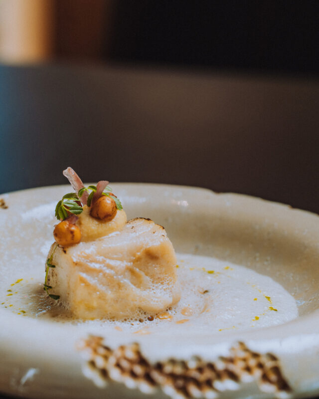 Delicately plated fish dish garnished with herbs and nuts, served on a textured white plate.