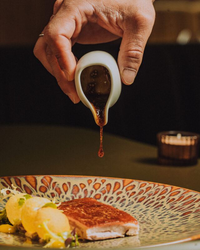 Hand pouring sauce from a small white vessel over a plated dish featuring a piece of meat and garnishes.
