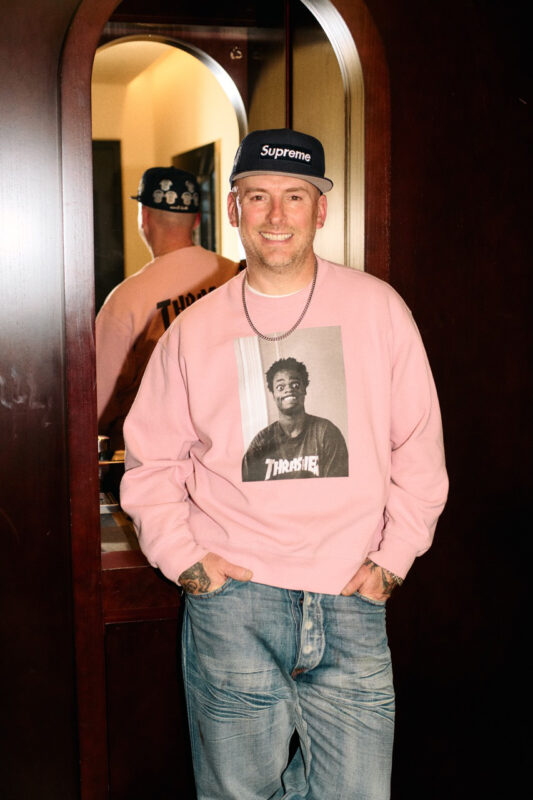 Man in a pink sweatshirt with a graphic print, wearing a Supreme cap, stands in front of a mirror, smiling.