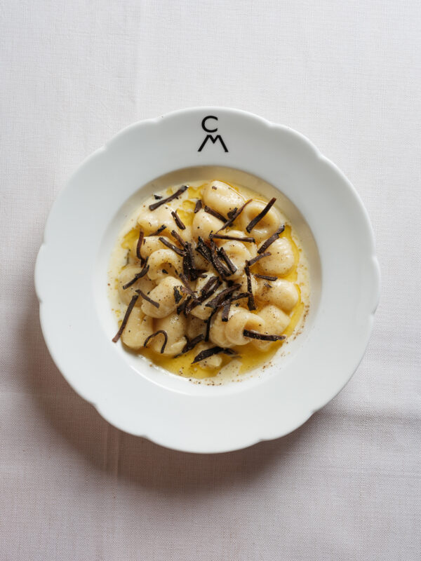 Gnocchi topped with brown butter and truffle shavings, served in a white bowl with a logo.