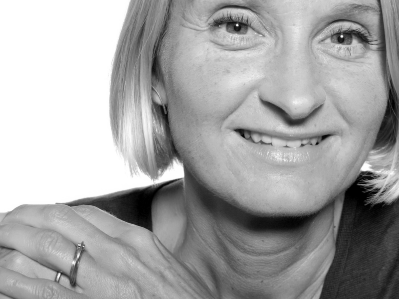 Close-up portrait of a smiling woman with short blonde hair, wearing a ring, against a bright white background.