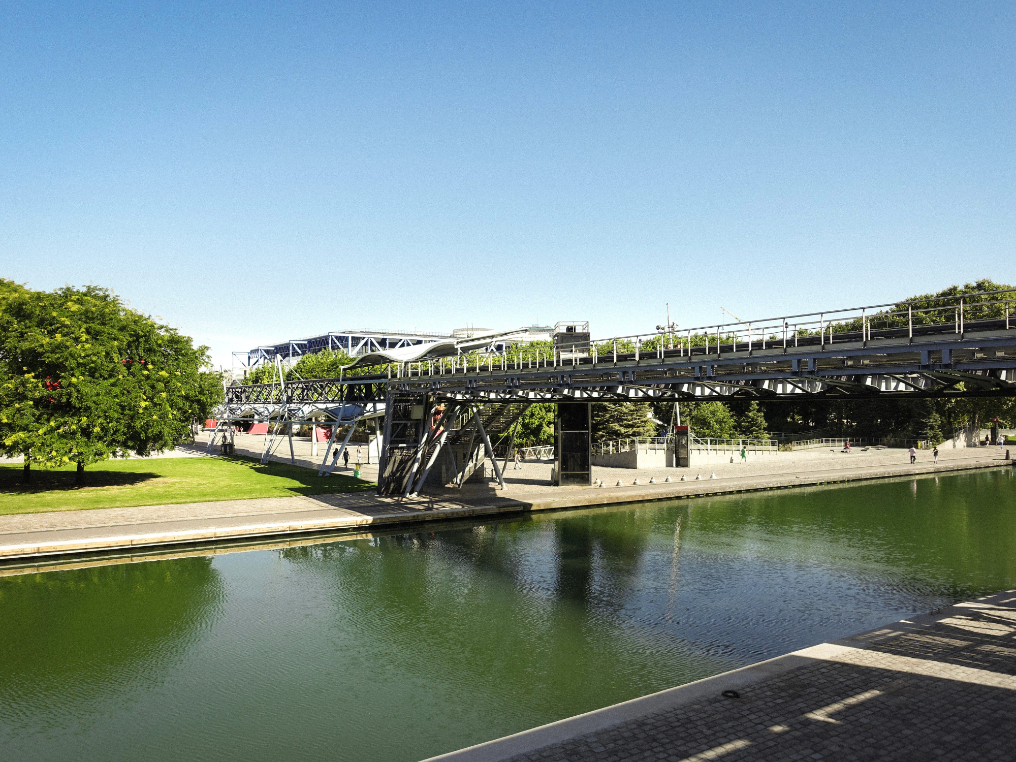A view of a modern pedestrian bridge over a calm waterway, flanked by greenery and urban architecture under a clear blue sky.