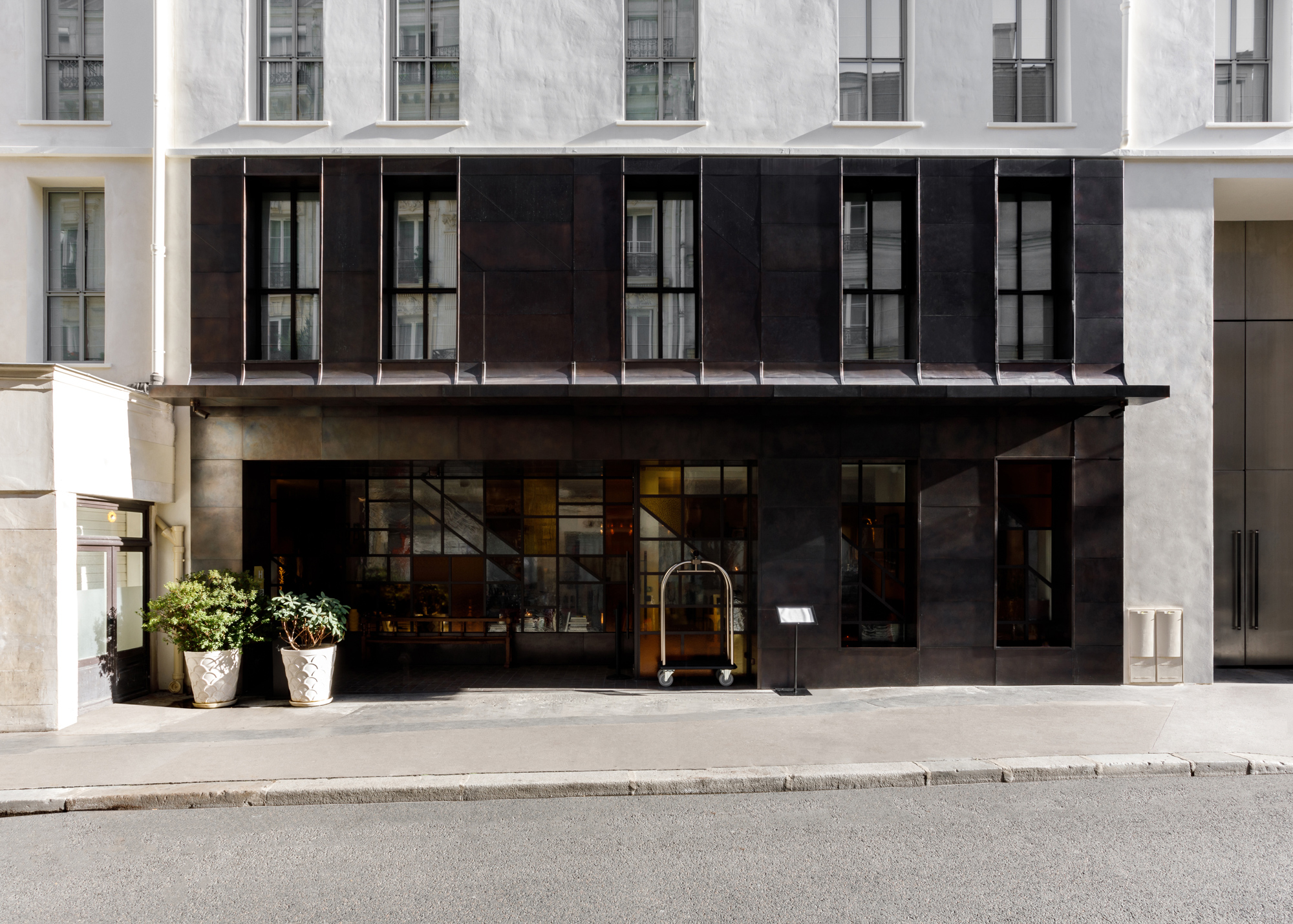 Modern black facade of Experimental Marais with large windows, potted plants, and a hotel cart outside.