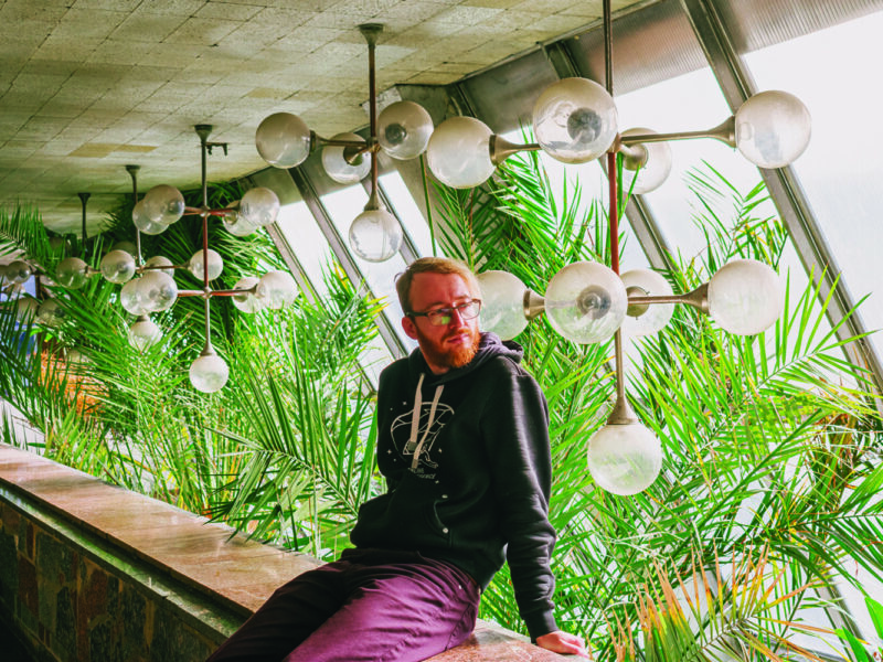 Man with glasses sitting on a ledge among large green plants and modern light fixtures in a bright space.