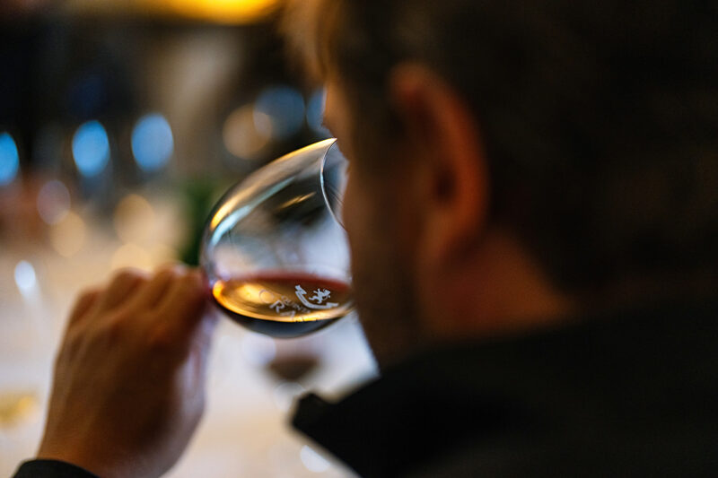 Person holding a wine glass, observing the wine's color, with blurred background elements suggesting a tasting event.