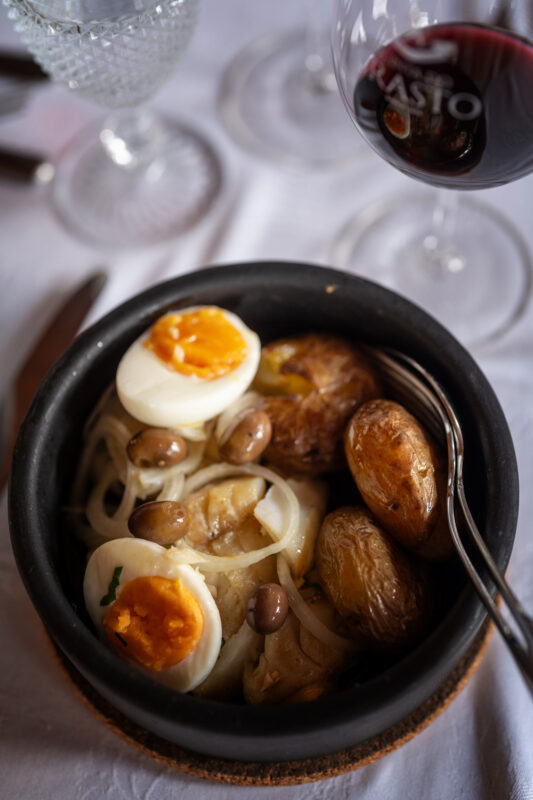 Traditional Portuguese dish featuring boiled eggs, potatoes, and olives in a black bowl, with red wine and glassware in th...