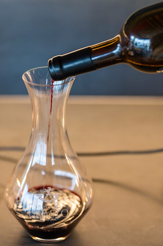 Red wine being poured from a bottle into a glass decanter on a countertop, with a blurred background.