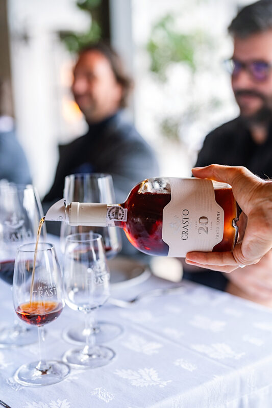 Hand pouring a bottle of Quinta do Crasto wine into a glass, with blurred figures of people in the background.