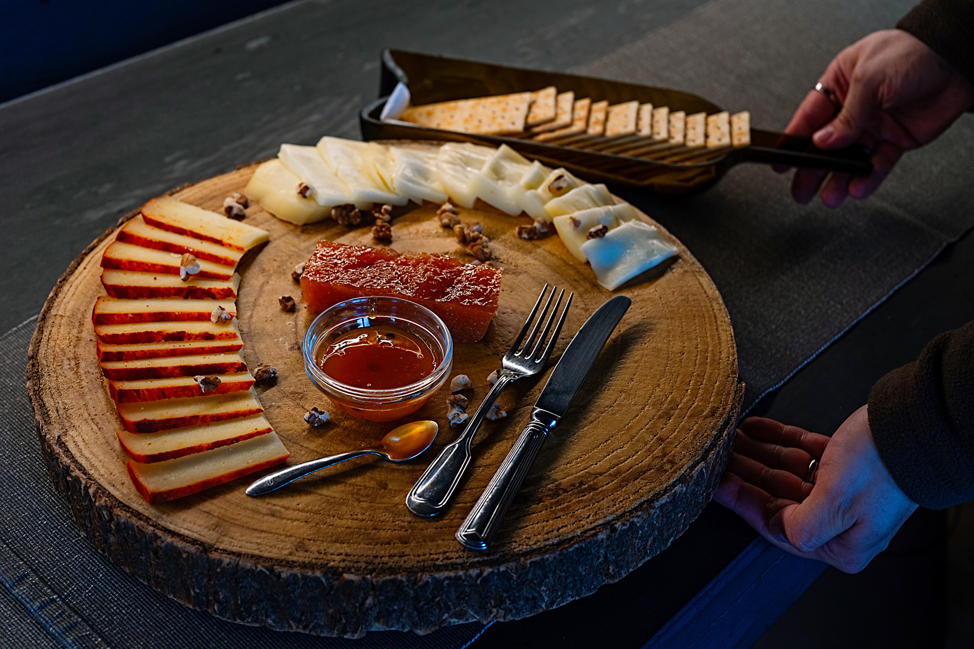 Cheese platter featuring assorted cheeses, honey, nuts, and crackers on a wooden board, with hands in the background.