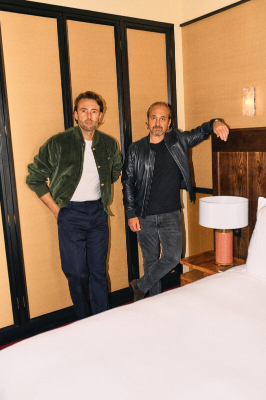 Two men pose in a stylish hotel room, one in a green jacket and the other in a black leather jacket, beside a bed.