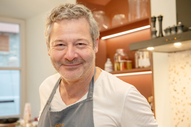 Smiling chef in a kitchen, wearing a gray apron, with shelves of ingredients and utensils in the background.