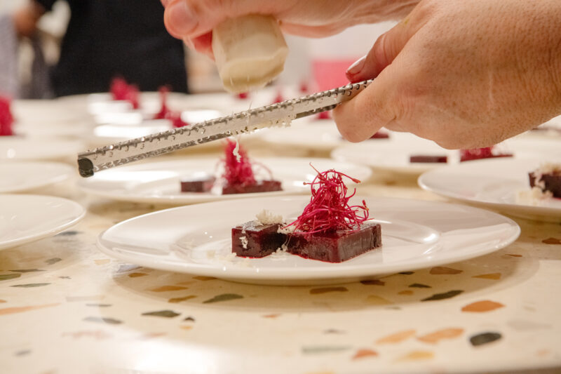 A hand grates cheese over a plated dish featuring beetroot and garnishes, with multiple plates in the background.