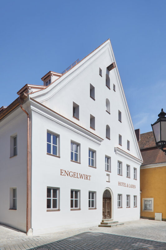 White building with a distinctive sloped roof, featuring large windows and the text "ENGELWIRT HOTEL & LÄDEN" on the facade.