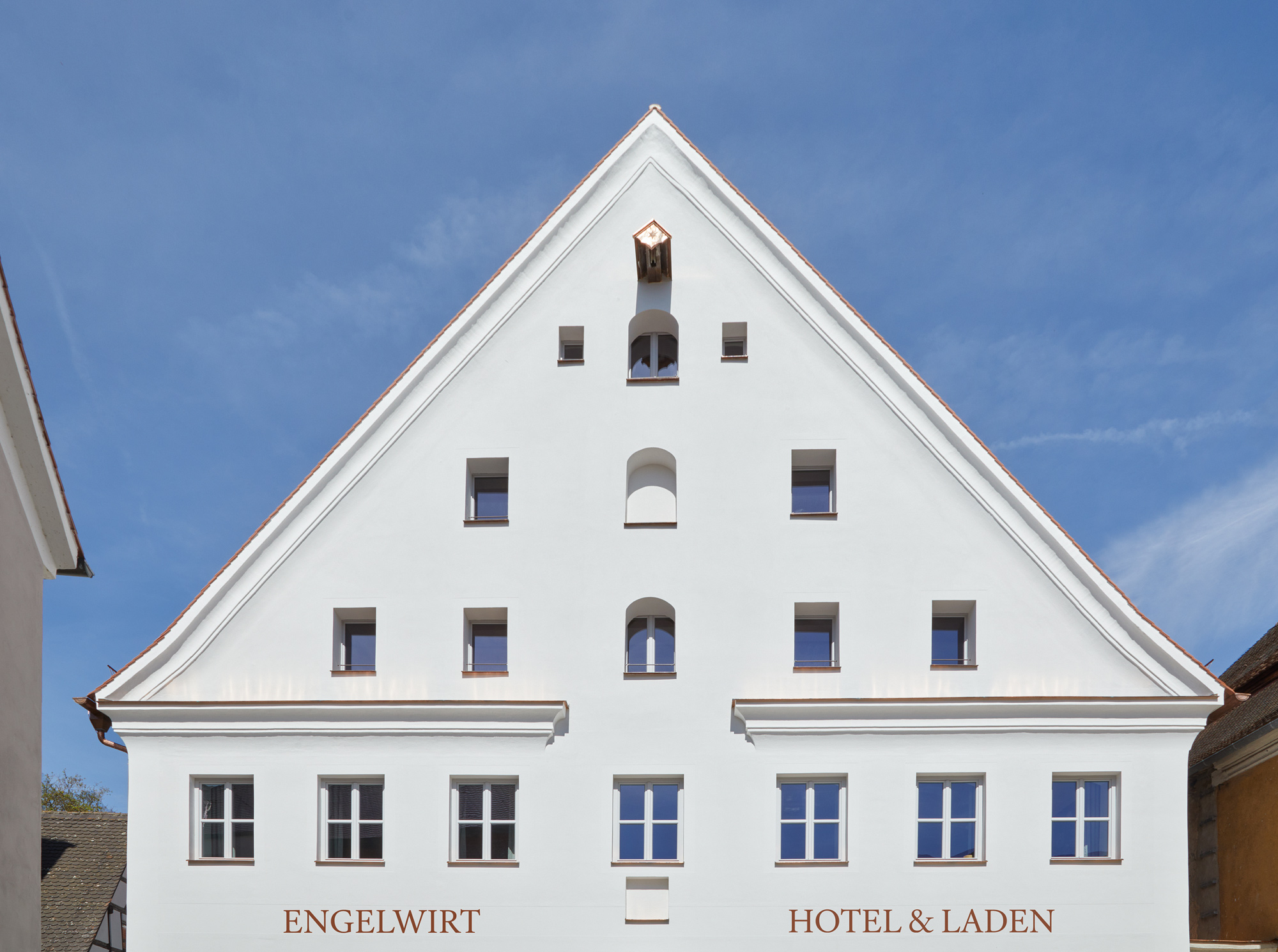 White triangular building facade with multiple windows, featuring "ENGELWIRT HOTEL & LADEN" signage. Clear blue sky above.