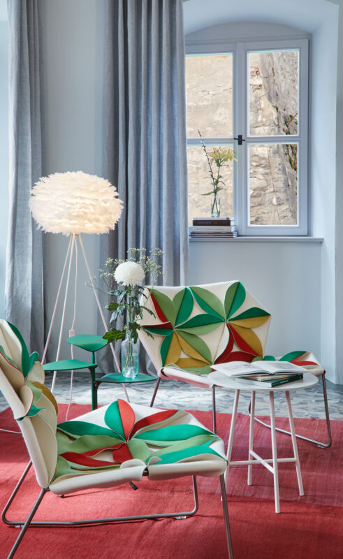 Colorful patterned chairs and a small table on a red rug, with a floor lamp and window in a stylish apartment setting.