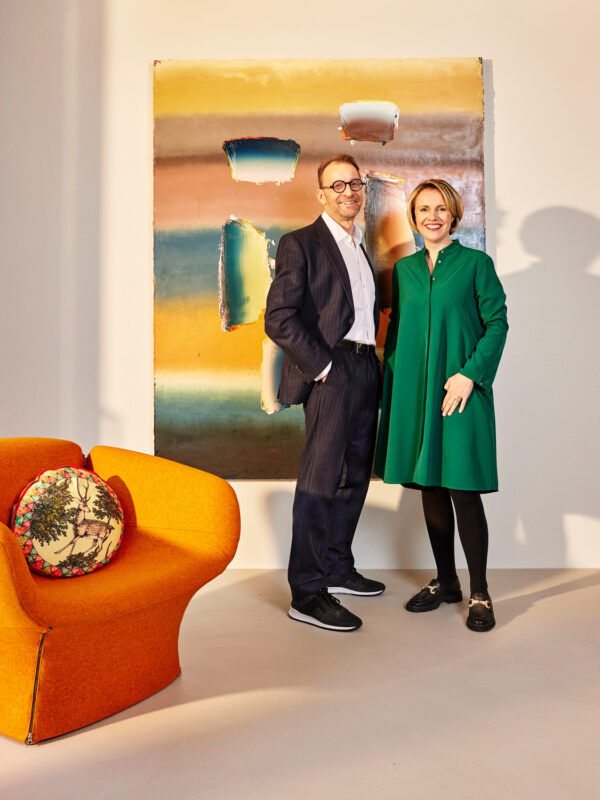 Couple standing beside a colorful abstract painting, with an orange chair and decorative pillow in the foreground.