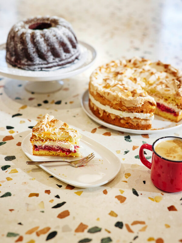 Layered cake with cream and almonds, slice on a plate, alongside a bundt cake and a cup of coffee on a speckled table.