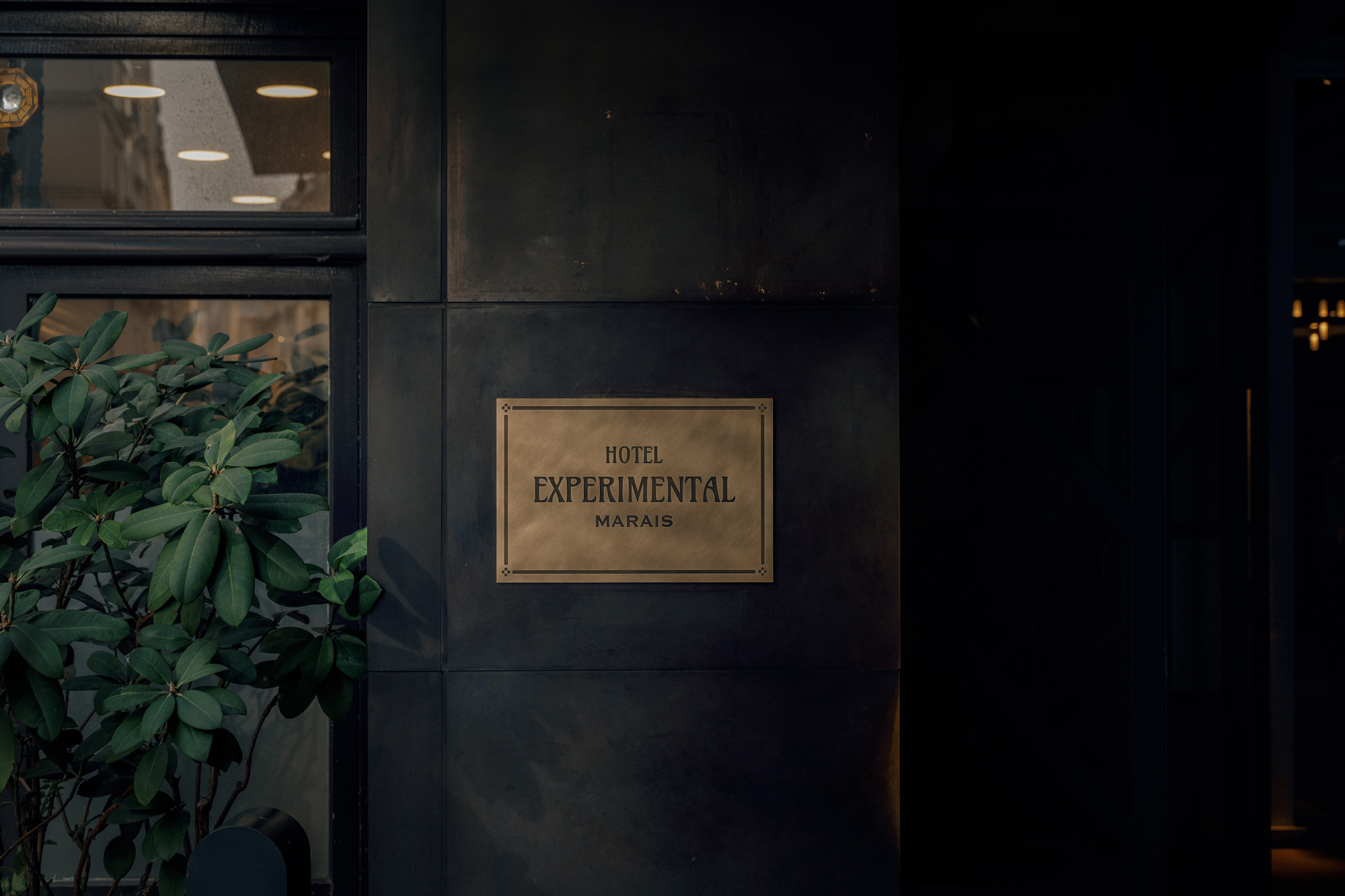 Signage for Hotel Experimental Marais, featuring a bronze plaque mounted on a dark wall beside a green plant.