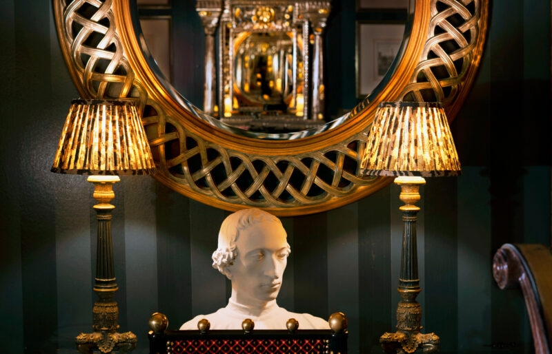 Bust of a man beneath an ornate mirror, flanked by two decorative lamps in a richly styled interior.